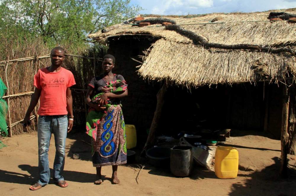 Lahima and Maxwell in front of their home. The thatch roof indicates that Maxwell has invested in assets before improving his home.